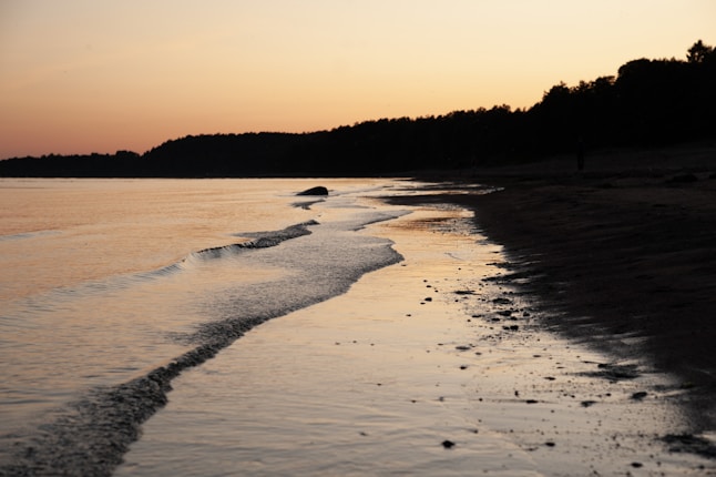 A peaceful shoreline at sunset with gentle waves lapping against the sandy beach. The sky is a gradient of warm orange and pink hues, fading into dusk. A silhouette of trees lines the horizon.