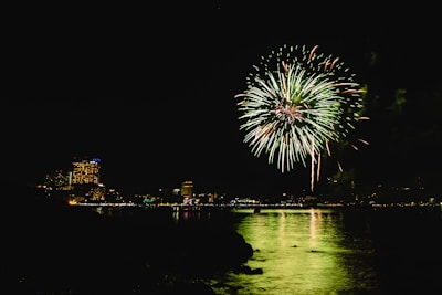 Bright green fireworks explode in the night sky over a calm body of water, reflecting the lights on its surface. In the distance, the skyline is illuminated by the colorful lights of buildings, adding to the festive atmosphere.