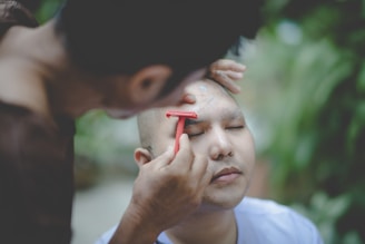 A person is carefully shaving another person's eyebrows with a red razor. The person receiving the shave has their eyes closed, appearing relaxed. The scene is outdoors, with a blurred background of greenery that provides a sense of calm and natural setting.