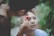 A person is carefully shaving another person's eyebrows with a red razor. The person receiving the shave has their eyes closed, appearing relaxed. The scene is outdoors, with a blurred background of greenery that provides a sense of calm and natural setting.