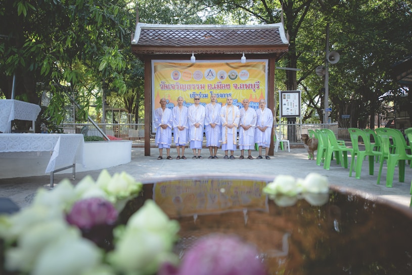 A group of people stands in front of a decorative banner in an outdoor setting, all dressed in similar robes. They are positioned on a paved area surrounded by green plastic chairs and lush trees. In the foreground, out-of-focus flowers float in a pond or water feature.