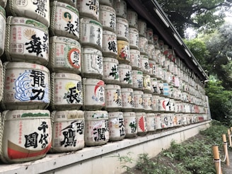 A long row of stacked traditional Japanese sake barrels, each adorned with colorful Japanese characters and symbols. The barrels are arranged in multiple tiers against a backdrop of lush, green foliage, creating a vibrant and cultural display.