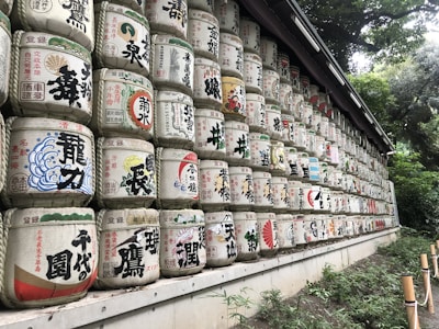 A long row of stacked traditional Japanese sake barrels, each adorned with colorful Japanese characters and symbols. The barrels are arranged in multiple tiers against a backdrop of lush, green foliage, creating a vibrant and cultural display.