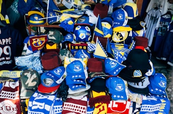 A vibrant display of sports merchandise with various hats, caps, and flags in blue and yellow, prominently featuring the Bosnia and Herzegovina flag and football-related items. There are shirts and scarves with team logos, including a jersey with the name Mbappe and the number 29.