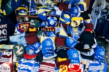 A vibrant display of sports merchandise with various hats, caps, and flags in blue and yellow, prominently featuring the Bosnia and Herzegovina flag and football-related items. There are shirts and scarves with team logos, including a jersey with the name Mbappe and the number 29.