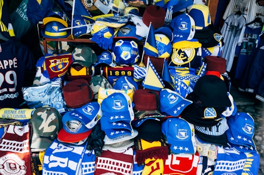 A vibrant display of sports merchandise with various hats, caps, and flags in blue and yellow, prominently featuring the Bosnia and Herzegovina flag and football-related items. There are shirts and scarves with team logos, including a jersey with the name Mbappe and the number 29.
