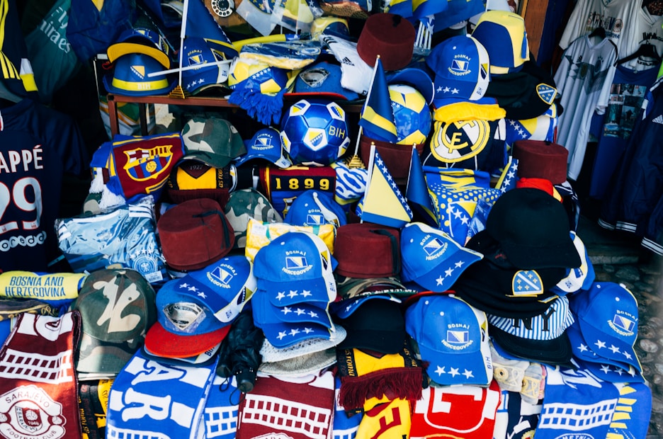 A vibrant display of sports merchandise with various hats, caps, and flags in blue and yellow, prominently featuring the Bosnia and Herzegovina flag and football-related items. There are shirts and scarves with team logos, including a jersey with the name Mbappe and the number 29.