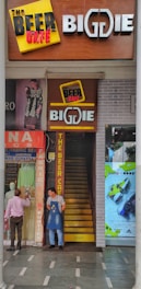 A narrow entrance with a staircase leads into a store named 'The Beer Cafe' and 'BigDIE'. Two men stand near the entrance, one wearing an apron. Above the stairway, there are signs and promotions related to beer. On the left side, a shop displays traditional clothing with mannequins and fabrics. On the right, an advertisement poster for sports shoes is visible, featuring bright colors.