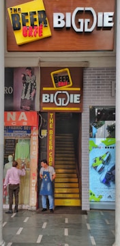 A narrow entrance with a staircase leads into a store named 'The Beer Cafe' and 'BigDIE'. Two men stand near the entrance, one wearing an apron. Above the stairway, there are signs and promotions related to beer. On the left side, a shop displays traditional clothing with mannequins and fabrics. On the right, an advertisement poster for sports shoes is visible, featuring bright colors.
