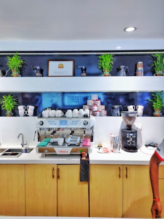 A coffee station with an espresso machine at the center, surrounded by coffee cups and a grinder on the right. Various coffee-related items and small potted plants are on the shelves above, along with a framed award marked 'Superbrand 2019'. The countertop is clean, with a sink on one side and cabinets underneath.