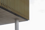 A modern architectural structure with vertical slats or louvers overhanging the side. The underside of the structure is supported by a cylindrical concrete column. The horizontal concrete slab and the vertical slats create a geometric pattern.