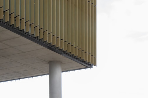 A modern architectural structure with vertical slats or louvers overhanging the side. The underside of the structure is supported by a cylindrical concrete column. The horizontal concrete slab and the vertical slats create a geometric pattern.