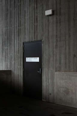 A dark, industrial-style fire safety door is set into a textured concrete wall. The door is equipped with a simple handle and features a sign that states 'Fire Safety Door - Do Not Obstruct - Do Not Keep Open.' Above the door, a small white box is mounted on the wall, possibly an alarm or sensor.
