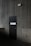 A dark, industrial-style fire safety door is set into a textured concrete wall. The door is equipped with a simple handle and features a sign that states 'Fire Safety Door - Do Not Obstruct - Do Not Keep Open.' Above the door, a small white box is mounted on the wall, possibly an alarm or sensor.