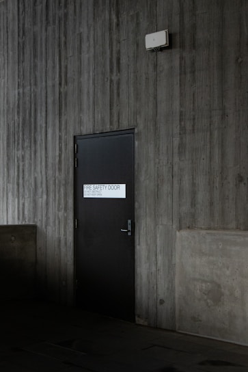 A dark, industrial-style fire safety door is set into a textured concrete wall. The door is equipped with a simple handle and features a sign that states 'Fire Safety Door - Do Not Obstruct - Do Not Keep Open.' Above the door, a small white box is mounted on the wall, possibly an alarm or sensor.