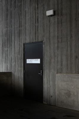 A dark, industrial-style fire safety door is set into a textured concrete wall. The door is equipped with a simple handle and features a sign that states 'Fire Safety Door - Do Not Obstruct - Do Not Keep Open.' Above the door, a small white box is mounted on the wall, possibly an alarm or sensor.