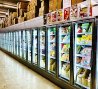 A row of brightly lit commercial refrigerators filled with various packaged foods, predominantly frozen goods. The tops of the refrigerators display a variety of cardboard boxes and posters featuring different products and individuals. The scene is set in a spacious supermarket or grocery store aisle with a reflective tile floor.