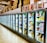 A row of brightly lit commercial refrigerators filled with various packaged foods, predominantly frozen goods. The tops of the refrigerators display a variety of cardboard boxes and posters featuring different products and individuals. The scene is set in a spacious supermarket or grocery store aisle with a reflective tile floor.