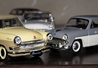 Three vintage model cars are positioned closely together, with a yellow car in the foreground, a grey car to its right, and another car partially visible in the background. The shiny details on the cars, such as the chrome grills and outlines, reflect light. The surface they rest on is smooth and reflective, contributing to the vintage aesthetic.