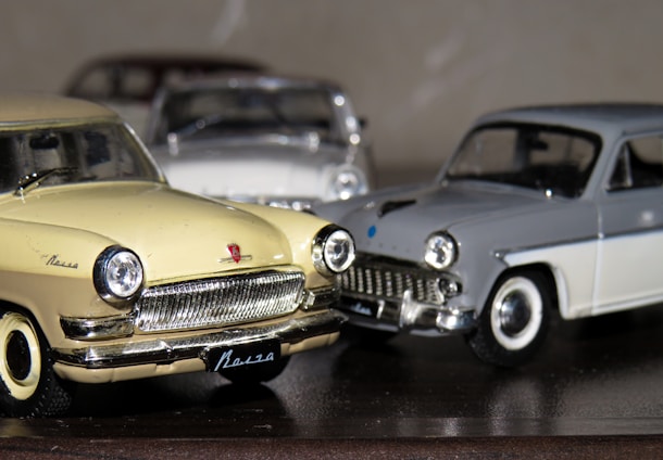 Three vintage model cars are positioned closely together, with a yellow car in the foreground, a grey car to its right, and another car partially visible in the background. The shiny details on the cars, such as the chrome grills and outlines, reflect light. The surface they rest on is smooth and reflective, contributing to the vintage aesthetic.