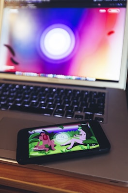 A smartphone displaying an app or game with cartoonish characters and greenery is resting on a laptop keyboard. The laptop screen shows a blurred, colorful background with a circular design.