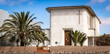 A church building with a textured white facade featuring a prominent cross near the roof. It is surrounded by lush palm trees and a stone fence, with a clear blue sky in the background.