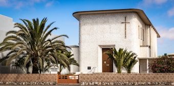 A church building with a textured white facade featuring a prominent cross near the roof. It is surrounded by lush palm trees and a stone fence, with a clear blue sky in the background.