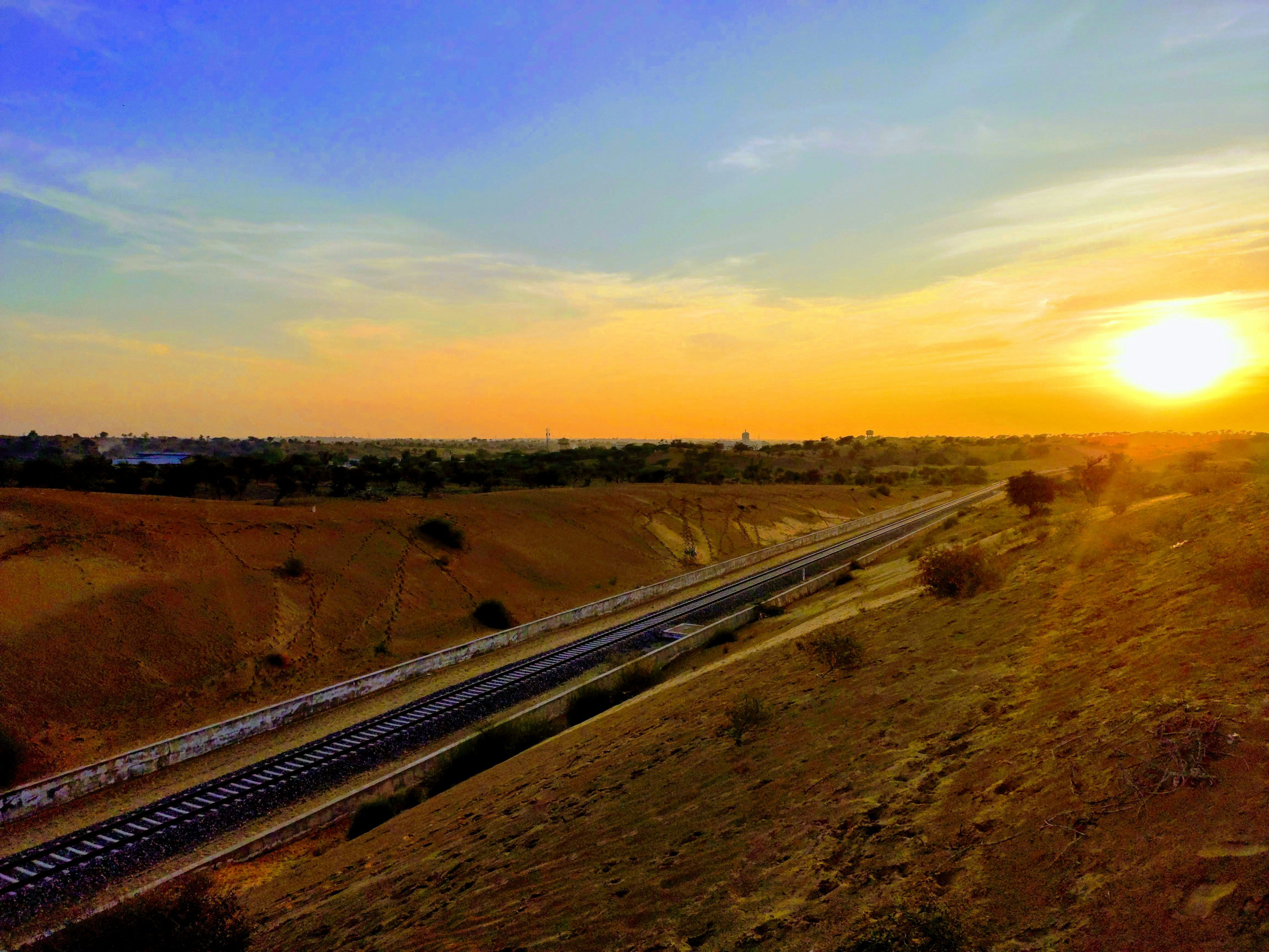 Freight train on long straight track through arid landscape at sunset, dramatic orange sky, long shadows, industrial rail corridor