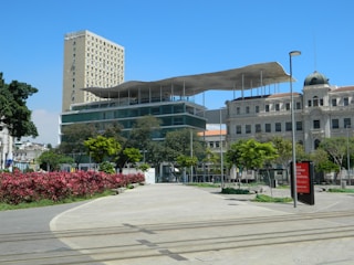 An urban scene featuring a modern structure with a unique floating roof design. The building is surrounded by greenery, including trees and pink flowering plants. In the background, there are other architectural buildings, one being a tall beige structure and another with a dome. The sky is clear and blue, and there are paths and tram tracks visible in the foreground.
