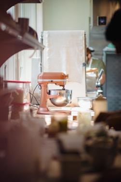 A kitchen scene featuring an orange standing mixer placed on a counter. Soft, natural light filters through a window, casting gentle shadows. In the background, a person wearing a cap and apron appears to be working, partially obscured by kitchen equipment and a towel.