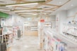 A well-lit, modern stationery store with neatly organized shelves filled with various stationery items. The ceiling features geometric wooden decorations and bright overhead lights. Merchandise includes pens, notebooks, and colorful packaging. Walls are decorated with minimalist art and signs, and there's an exit sign visible above one section.