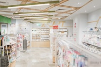 A well-lit, modern stationery store with neatly organized shelves filled with various stationery items. The ceiling features geometric wooden decorations and bright overhead lights. Merchandise includes pens, notebooks, and colorful packaging. Walls are decorated with minimalist art and signs, and there's an exit sign visible above one section.