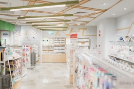 A well-lit, modern stationery store with neatly organized shelves filled with various stationery items. The ceiling features geometric wooden decorations and bright overhead lights. Merchandise includes pens, notebooks, and colorful packaging. Walls are decorated with minimalist art and signs, and there's an exit sign visible above one section.