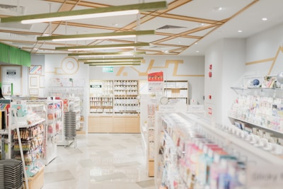 A well-lit, modern stationery store with neatly organized shelves filled with various stationery items. The ceiling features geometric wooden decorations and bright overhead lights. Merchandise includes pens, notebooks, and colorful packaging. Walls are decorated with minimalist art and signs, and there's an exit sign visible above one section.