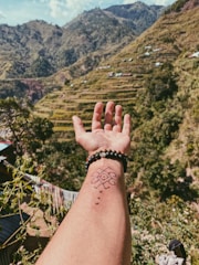 A hand with a tattoo and beaded bracelets is outstretched against a backdrop of lush, terraced mountains. The scene includes greenery, small structures on the hillsides, and a clear sky.