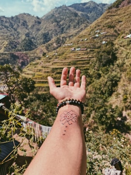 A hand with a tattoo and beaded bracelets is outstretched against a backdrop of lush, terraced mountains. The scene includes greenery, small structures on the hillsides, and a clear sky.