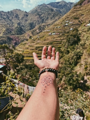A hand with a tattoo and beaded bracelets is outstretched against a backdrop of lush, terraced mountains. The scene includes greenery, small structures on the hillsides, and a clear sky.