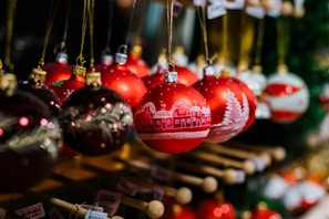 A collection of red and dark-colored Christmas ornaments decorated with white illustrations of snow-covered houses and trees, hanging from a rack. The ornaments are shiny, reflecting light, and are suspended by golden strings. The background appears softly blurred, suggesting a festive setting.