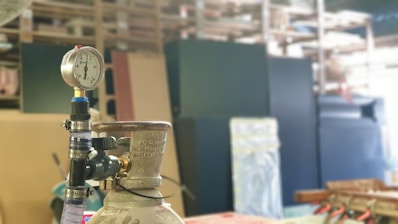 A pressure gauge mounted on top of a metallic cylinder is in focus. The background consists of blurry industrial shelving and equipment, which might include large black and beige panels.