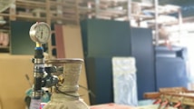 A pressure gauge mounted on top of a metallic cylinder is in focus. The background consists of blurry industrial shelving and equipment, which might include large black and beige panels.