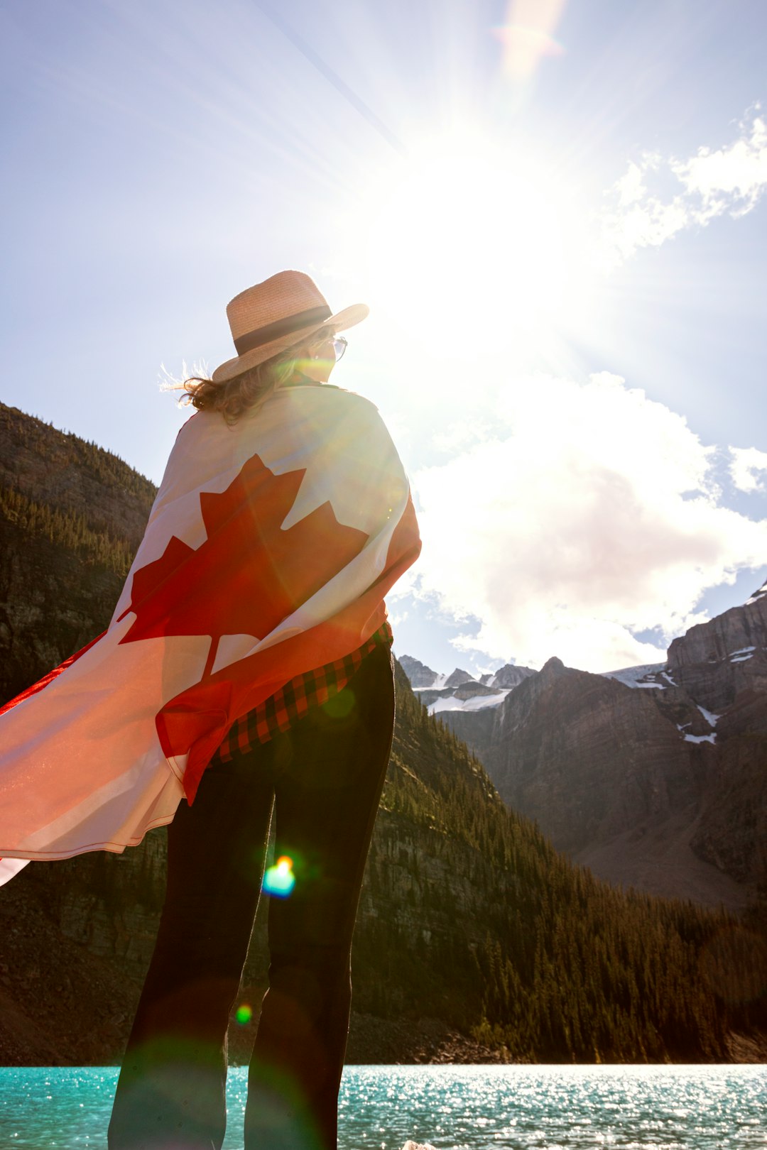 woman wearing black pants and white hat covering Canada flag while standing near body of water viewing mountain under white and blue sky