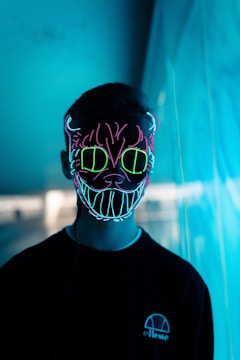 A person is wearing a glowing neon mask with intricate patterns and a devilish smile. The mask features bright neon colors including blue, pink, and green, and the background is illuminated with a blue hue, creating a moody and futuristic atmosphere. The person is dressed in a dark outfit, adding contrast to the vibrant mask.