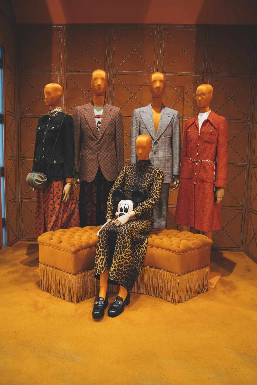 Quattro manichini che indossano abiti formali e un altro manichino femminile che indossa un abito a maniche lunghe leopardato marrone seduto su un pouf arancione trapuntato