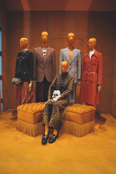 four mannequins wearing formal suits and another female mannequin wearing brown leopard long-sleeved dress sitting on tufted orange ottoman