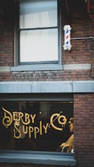 A vintage-style barbershop with brick walls and a large window displaying ornate gold lettering spelling 'Derby Supply Co'. A classic barber pole is mounted on the building's exterior. Inside, a barber is attending to a customer in a chair.