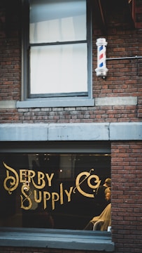 A vintage-style barbershop with brick walls and a large window displaying ornate gold lettering spelling 'Derby Supply Co'. A classic barber pole is mounted on the building's exterior. Inside, a barber is attending to a customer in a chair.