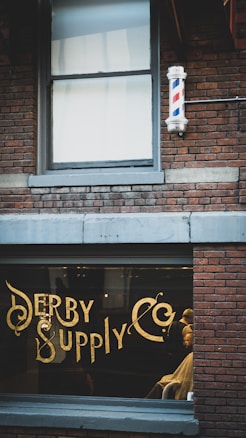 A vintage-style barbershop with brick walls and a large window displaying ornate gold lettering spelling 'Derby Supply Co'. A classic barber pole is mounted on the building's exterior. Inside, a barber is attending to a customer in a chair.