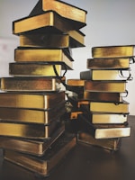Two stacks of small, compact, golden-edged books are arranged on a dark surface. The books have black ribbons attached, suggesting they may be used as bookmarks. The lighting accentuates the metallic sheen of the book edges, creating a reflective and warm appearance.