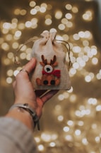A hand holding a small burlap gift bag adorned with a decorative reindeer applique is prominently featured. The background consists of numerous blurred warm yellow lights, creating a festive and cozy atmosphere.