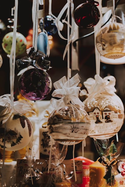 A collection of beautifully decorated Christmas ornaments is hanging, each with intricate designs. The ornaments showcase various festive scenes and patterns, adorned with ribbons and bows. Soft lighting enhances the warm and cozy atmosphere.